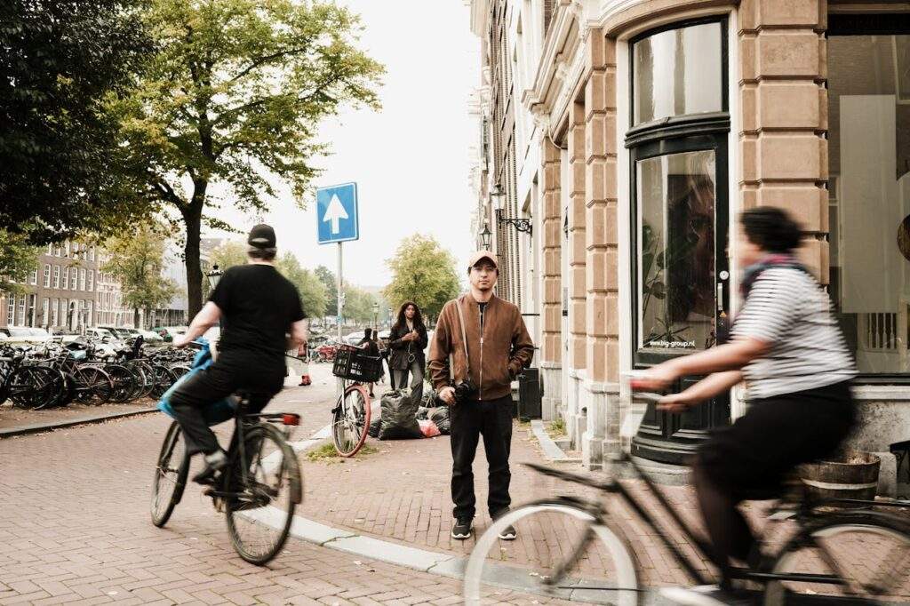 Man waiting to cross a street, people on bikes, and more bikes parked on the side.